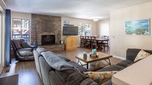 A living room with a stone fireplace, a television, a dining area with wooden furniture, and a large window. - Enjoy a Cottage near the lake 2bdr, Tahoe 2Bdr Postmasters Cottage (South Lake Tahoe)