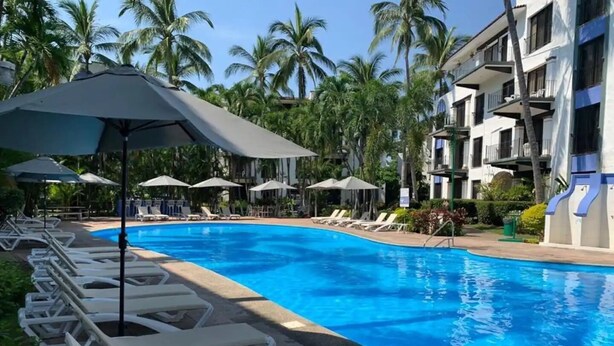 A swimming pool with white lounge chairs and umbrellas, surrounded by palm trees and a multi-story building. - CONDO BELLA BY TRVL2HM (Puerto Vallarta)