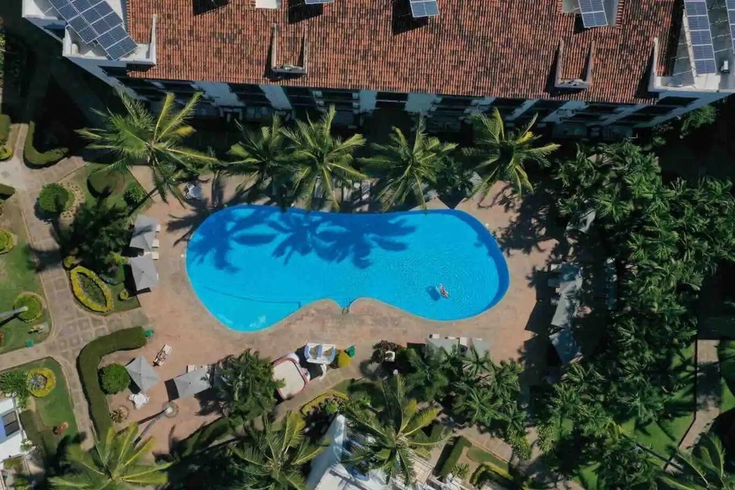 An aerial view of a hotel with a swimming pool surrounded by palm trees and a tiled roof.
