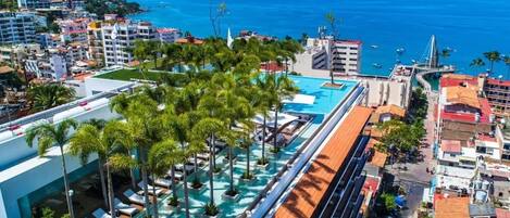 A rooftop pool with palm trees, a view of the sea, and surrounding buildings.