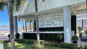 A modern outdoor bar area with a marble wall, surrounded by palm trees and greenery, overlooking a cityscape. - CONDO 57 BY TRVL2HM (Puerto Vallarta)