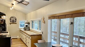 Cocina moderna con comedor, suelo de madera y ventanas amplias con vistas a árboles.