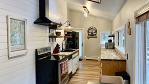A modern kitchen with a black stove, wooden cabinets, and a tiled backsplash. There is a dining area with a table and chairs, and a window providing natural light. - Modern Cozy Mountain Cabin with Private Hot Tub • Treehouse • Fire Pit (Lyons)