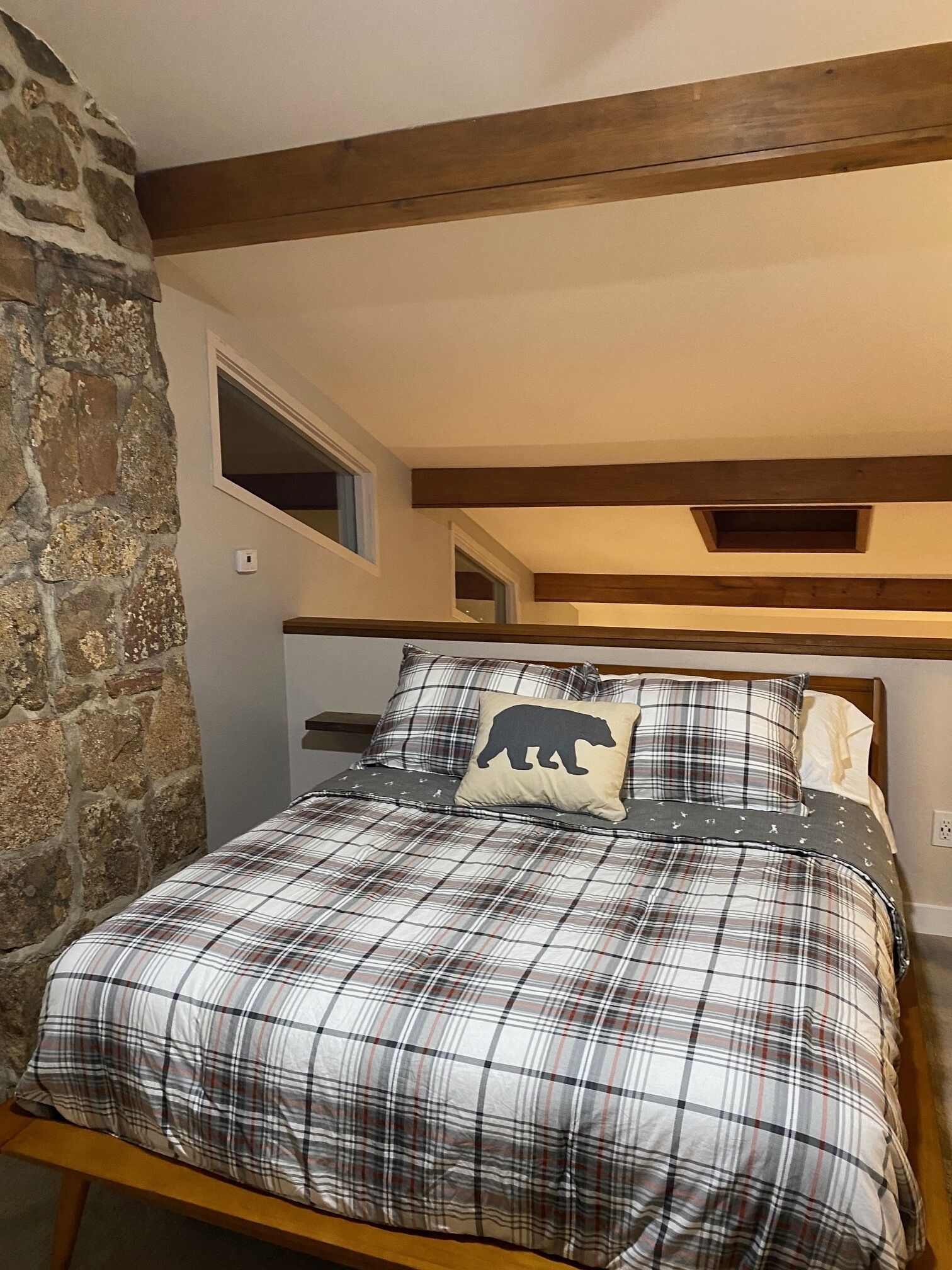 Une chambre avec une couvre-lit à carreaux, un coussin en forme d’ours, un mur en pierre et une poutre en bois au plafond.