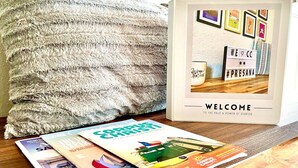 A magazine on a wooden table, a framed welcome sign, and a fluffy pillow.
