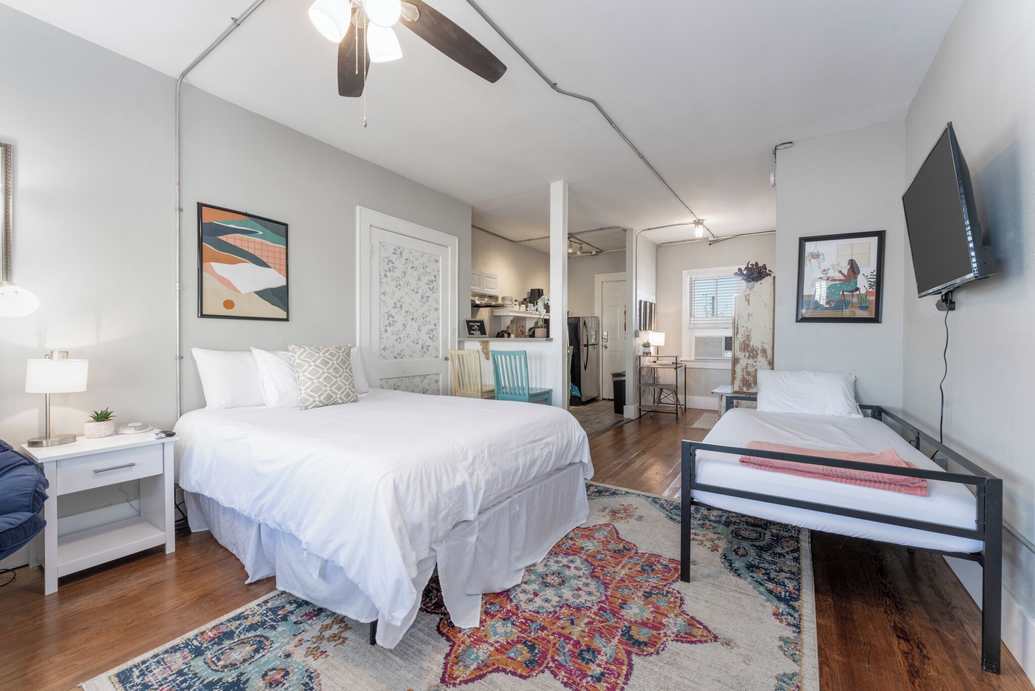 A bedroom with a bed, a desk, a chair, a TV, and a kitchen in the background.