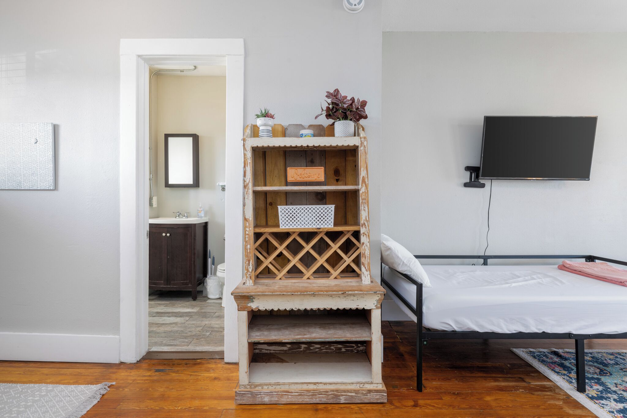 Een slaapkamer met een bed, een aan de muur bevestigde televisie, een houten plank met decoratieve objecten en een deur naar een badkamer.