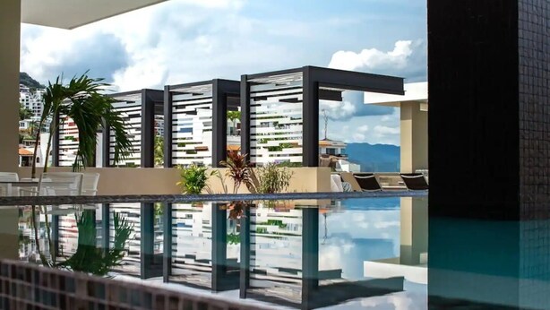 A modern pool area with clear reflections, lounge chairs, and a view of buildings and mountains. - CONDO L268  BY TRVL2HM (Puerto Vallarta)