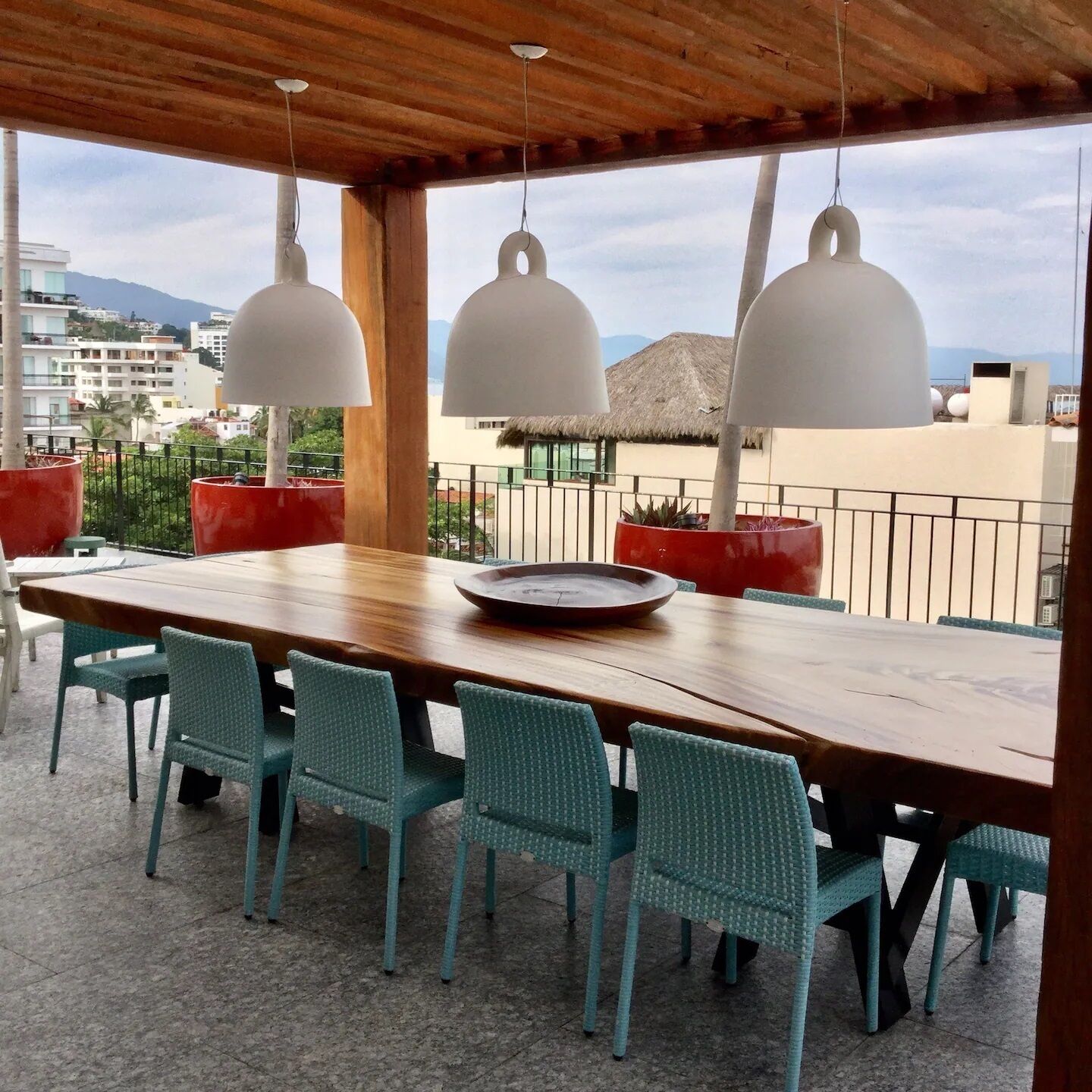 An outdoor dining area with a wooden table and blue chairs, three hanging pendant lights, and a view of buildings and mountains in the background.
