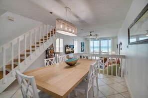A dining area with a wooden table, white chairs, a hanging light fixture, and a staircase with white railings.