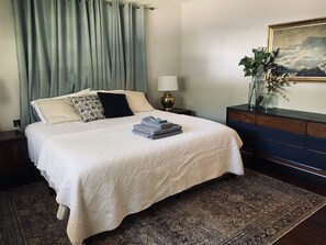 A neatly made bed with a white bedspread and pillows, a bedside table with a lamp, a dresser with a vase of plants, and a painting on the wall.