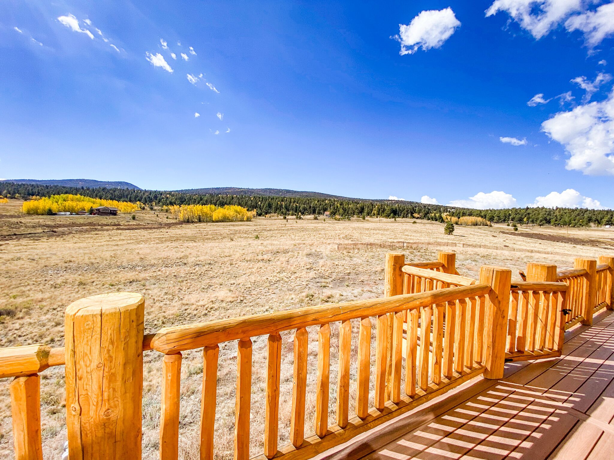 A wooden deck with a view of a dry grass field, trees, and a clear sky.