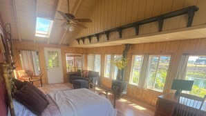 A spacious room with a large bed, a wooden ceiling, and a view of the outdoors through multiple windows.