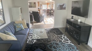 A living room with a sofa, coffee table, and TV stand. There is a dining area in the background with chairs and a table.