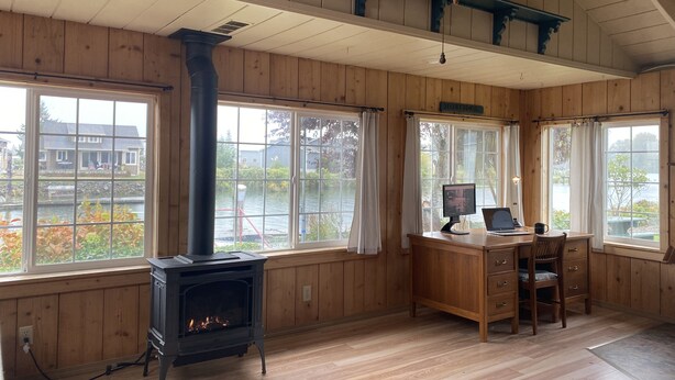 A room with wooden walls and a large window overlooking a body of water. A fireplace is built into the wall, and there is a desk with a computer and chair.