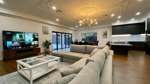 A modern living room with a large flat-screen TV, a beige sofa, a glass coffee table, and a kitchen area in the background. - New & Spacious Luxury Mansion with 5 Bedrooms & 4 (Unley)