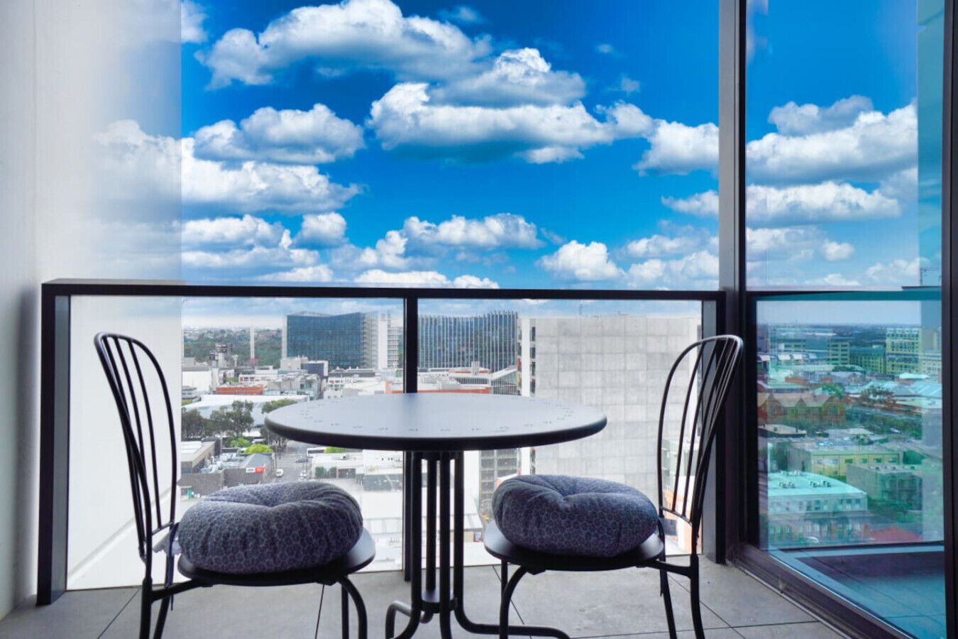 Un balcon avec une table ronde et deux chaises, une vue sur la ville à travers de grandes fenêtres et un ciel bleu dégagé parsemé de nuages.
