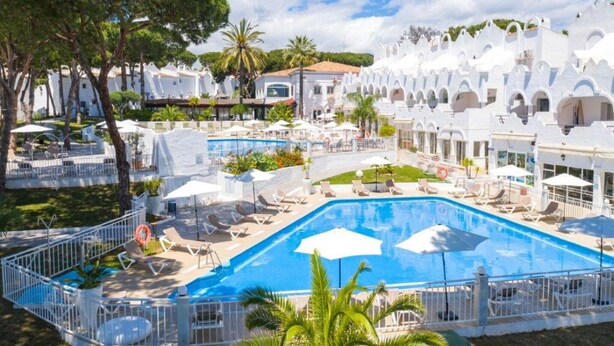 Un resort con una grande piscina, lettini, ombrelloni ed edifici bianchi con archi.