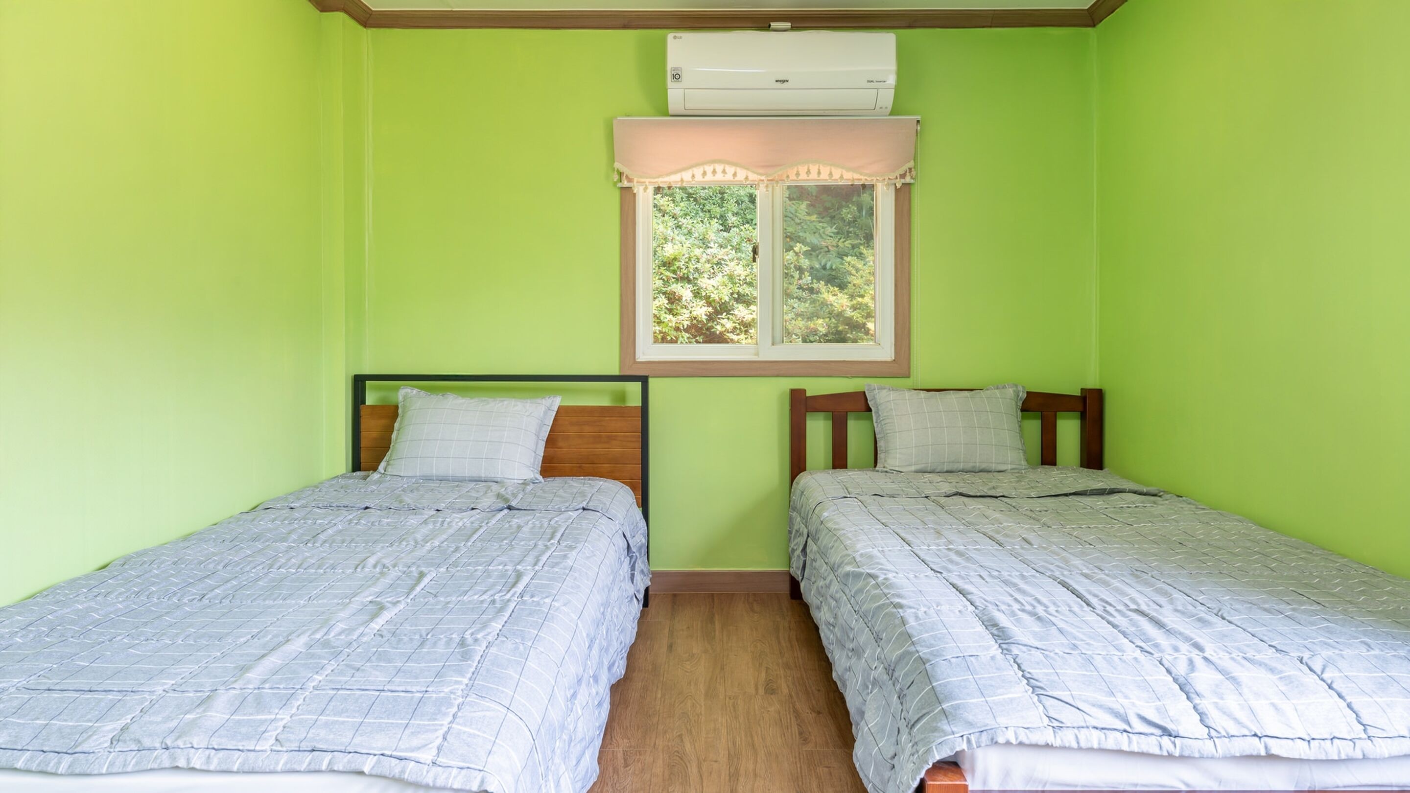 Une chambre avec deux lits, des murs verts, des têtes de lit en bois et une fenêtre donnant sur des arbres.