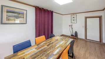 A room with a wooden table, orange and blue chairs, a red curtain, a framed picture, a door, and a small decorative tree.