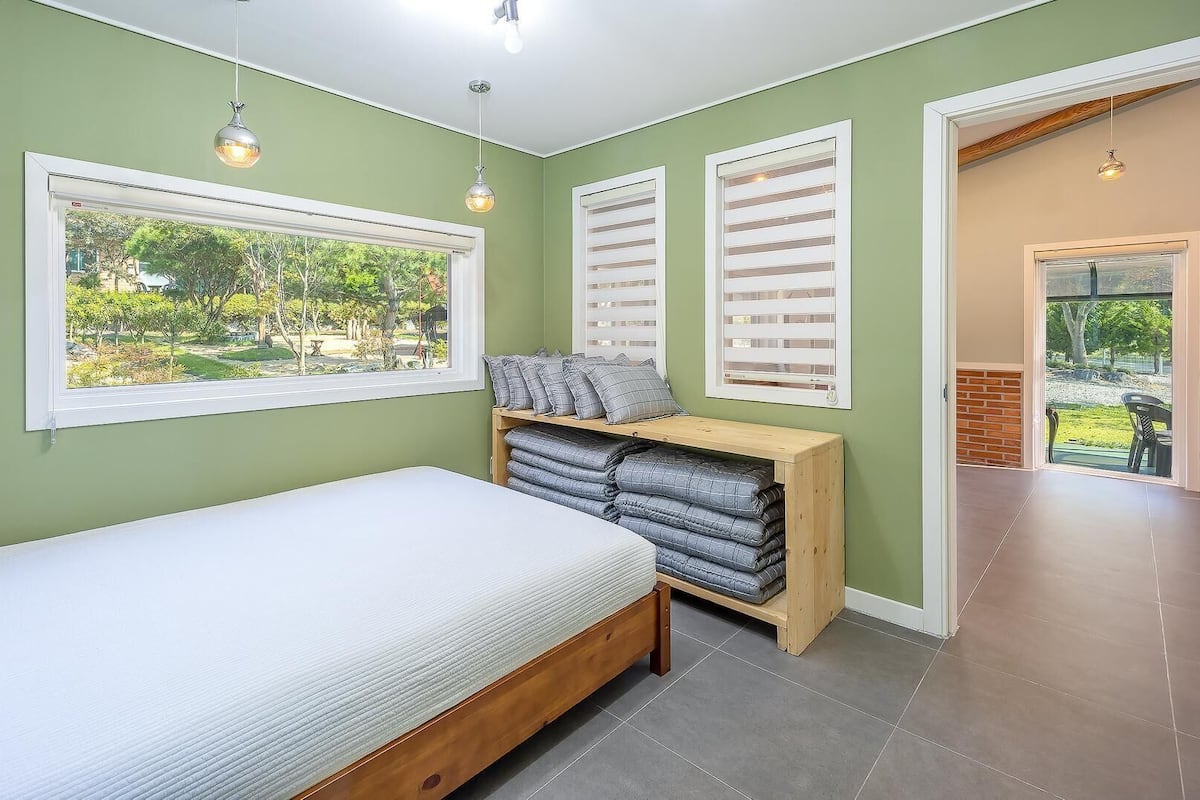 Un dormitorio con cama, un banco de madera con almohadas, una ventana con persianas y vistas al exterior.