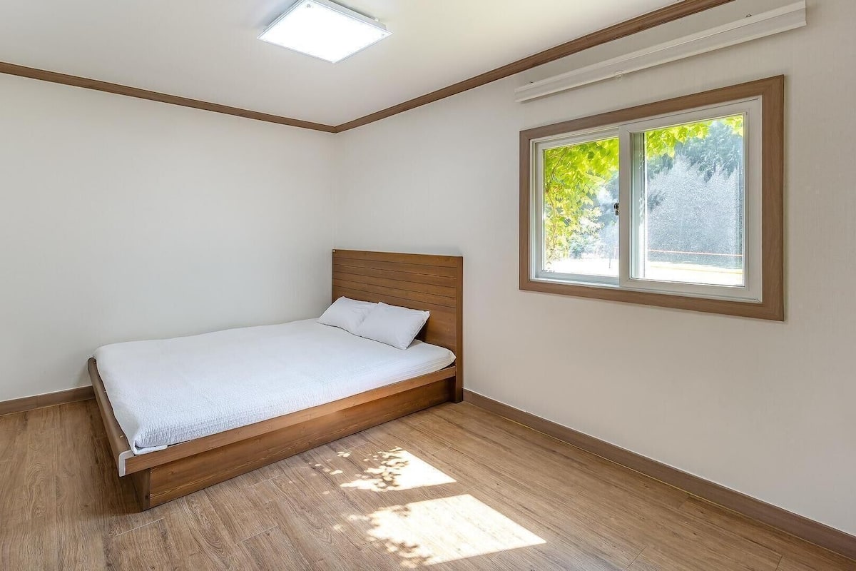Una cama individual en una habitación con techo inclinado, una ventana con vista a árboles y suelo de madera.