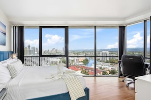 A hotel room with a large bed, a desk, and a view of a cityscape through large windows.