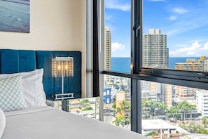 Una habitació d'hotel amb una finestra gran, un llit amb roba de llit blanca, un reposacapses blau fosc i una làmpada de peu decorativa.