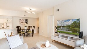 Un salon moderne avec un téléviseur à écran plat, un canapé, un coin repas avec des chaises et une cuisine en arrière-plan.