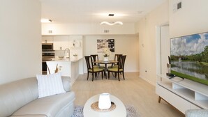 Un salon moderne avec un canapé, une table basse, un coin repas avec des chaises, une cuisine en arrière-plan et un téléviseur posé sur un meuble.