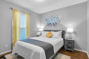 A bedroom with a bed, two bedside lamps, a nightstand, a window with yellow curtains, and three wall-mounted paintings.