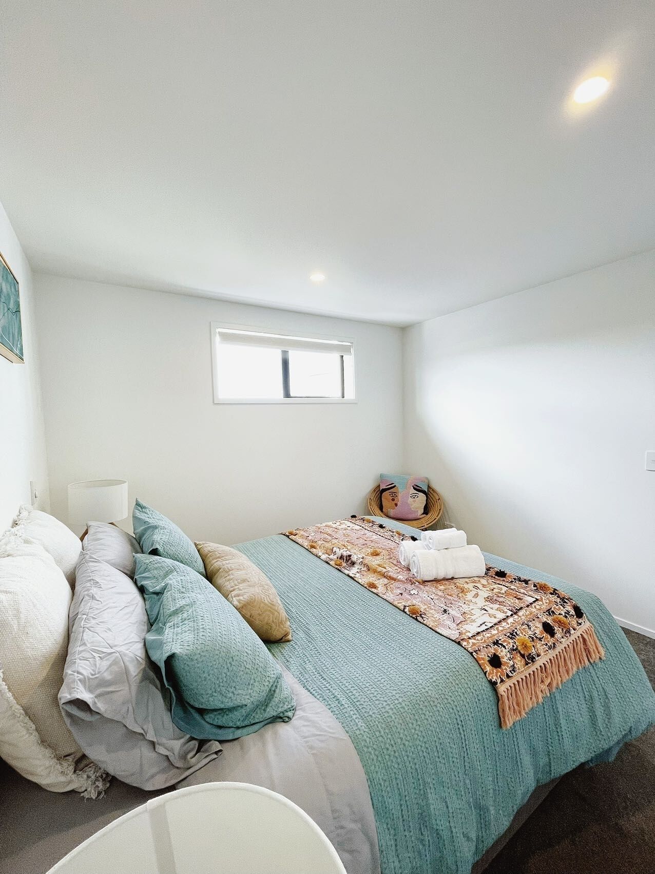 A bedroom with a bed, pillows, a blanket, a small window, and a wall-mounted lamp.