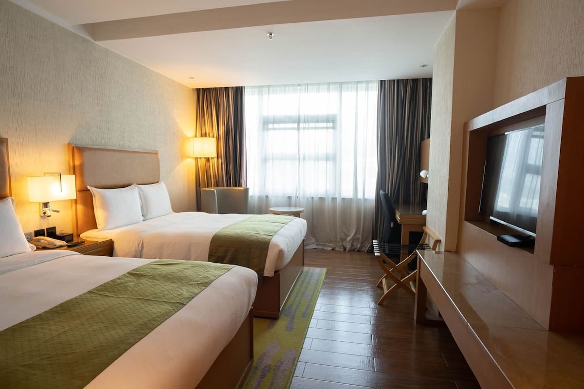 A hotel room with two beds, a desk, a chair, a TV, and a window with curtains.