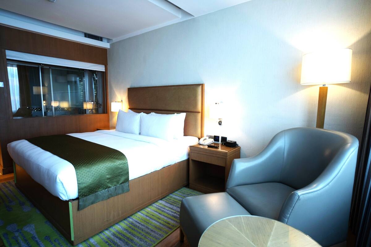 A hotel room with a large bed, a chair, a nightstand, and a window with blinds.