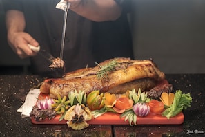 A chef carving a roasted pork leg on a red cutting board with various vegetables and herbs. - Ion Hotel (Baguio)