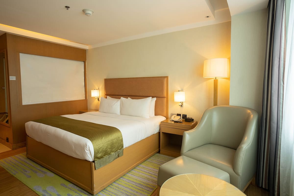 A hotel room with a bed, two bedside lamps, a chair, a small table, and a window with curtains.