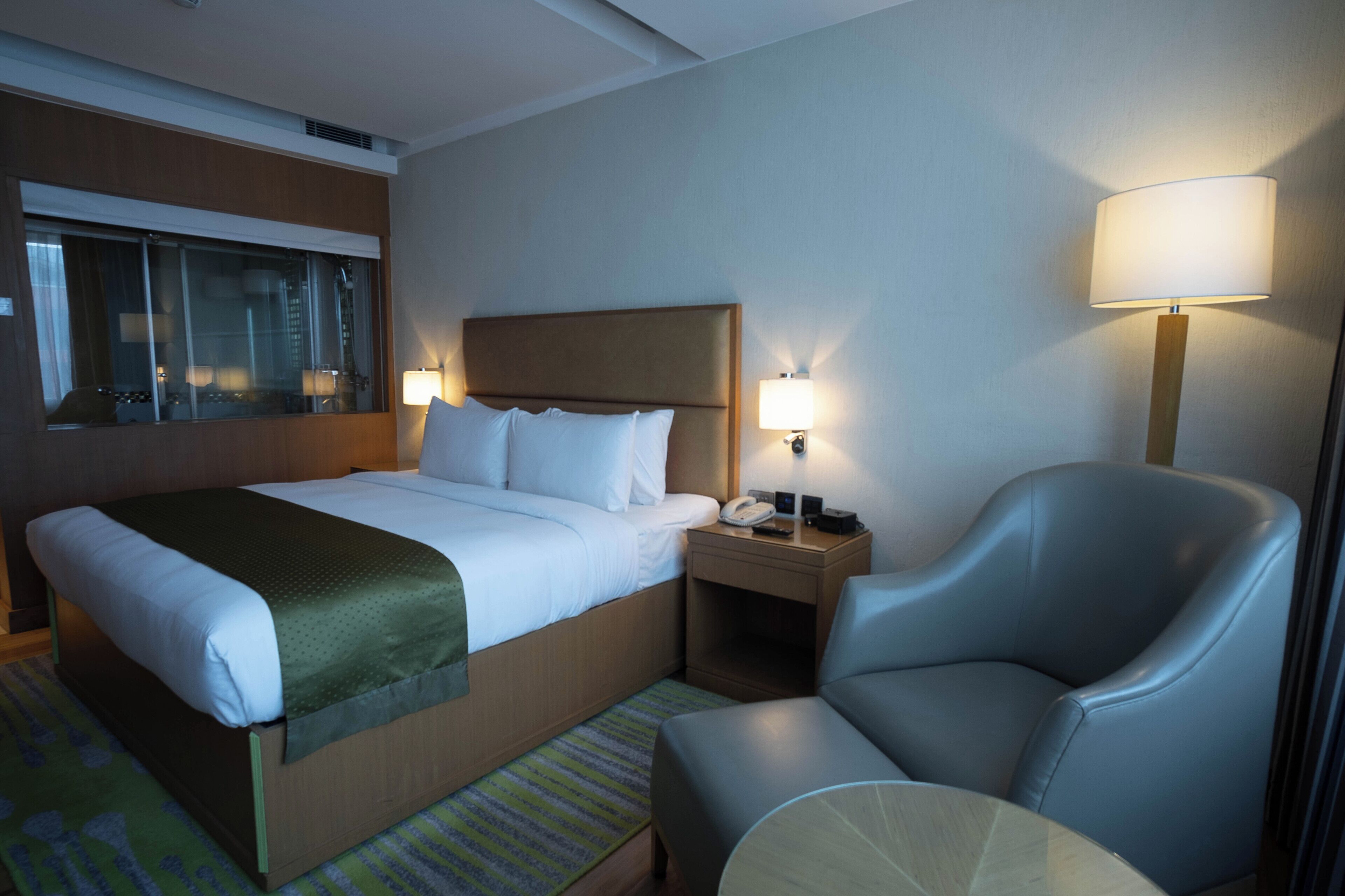 Een hotelkamer met een groot bed, een stoel, een nachtkastje met een lamp en een raam met gordijnen.
