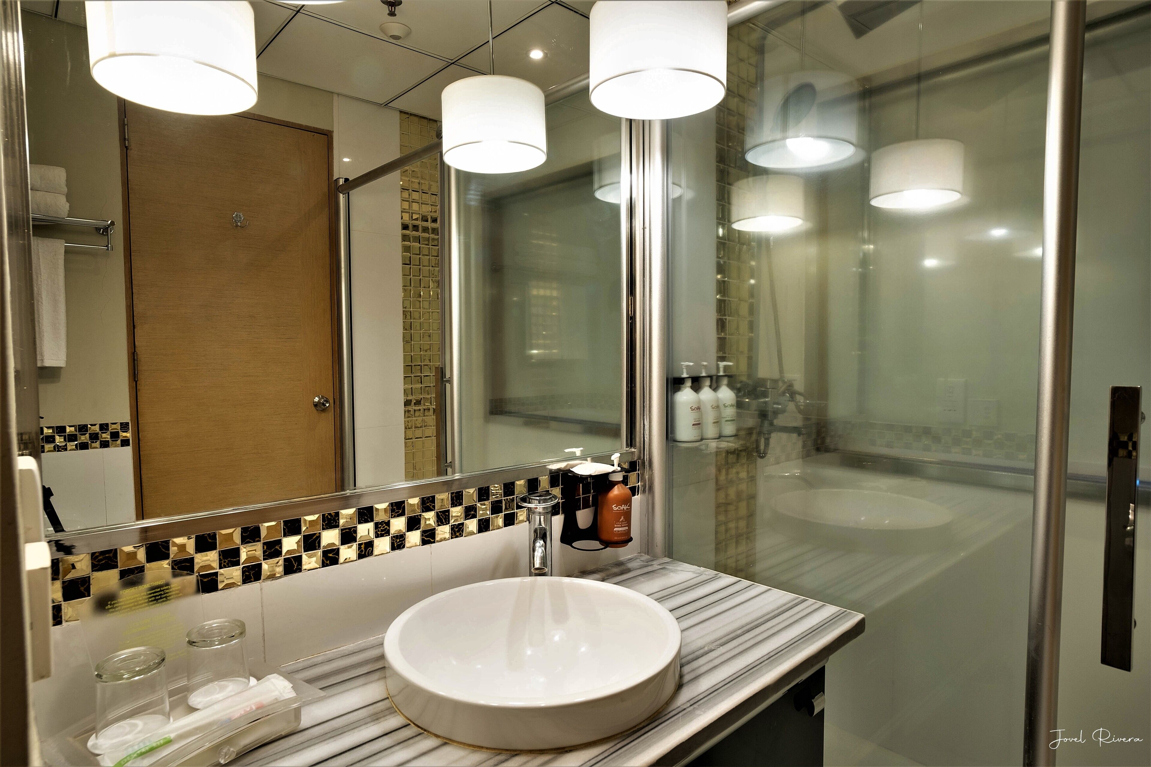 A modern bathroom with a marble sink, glass shower enclosure, and a mirror with a black and gold checkered backsplash.