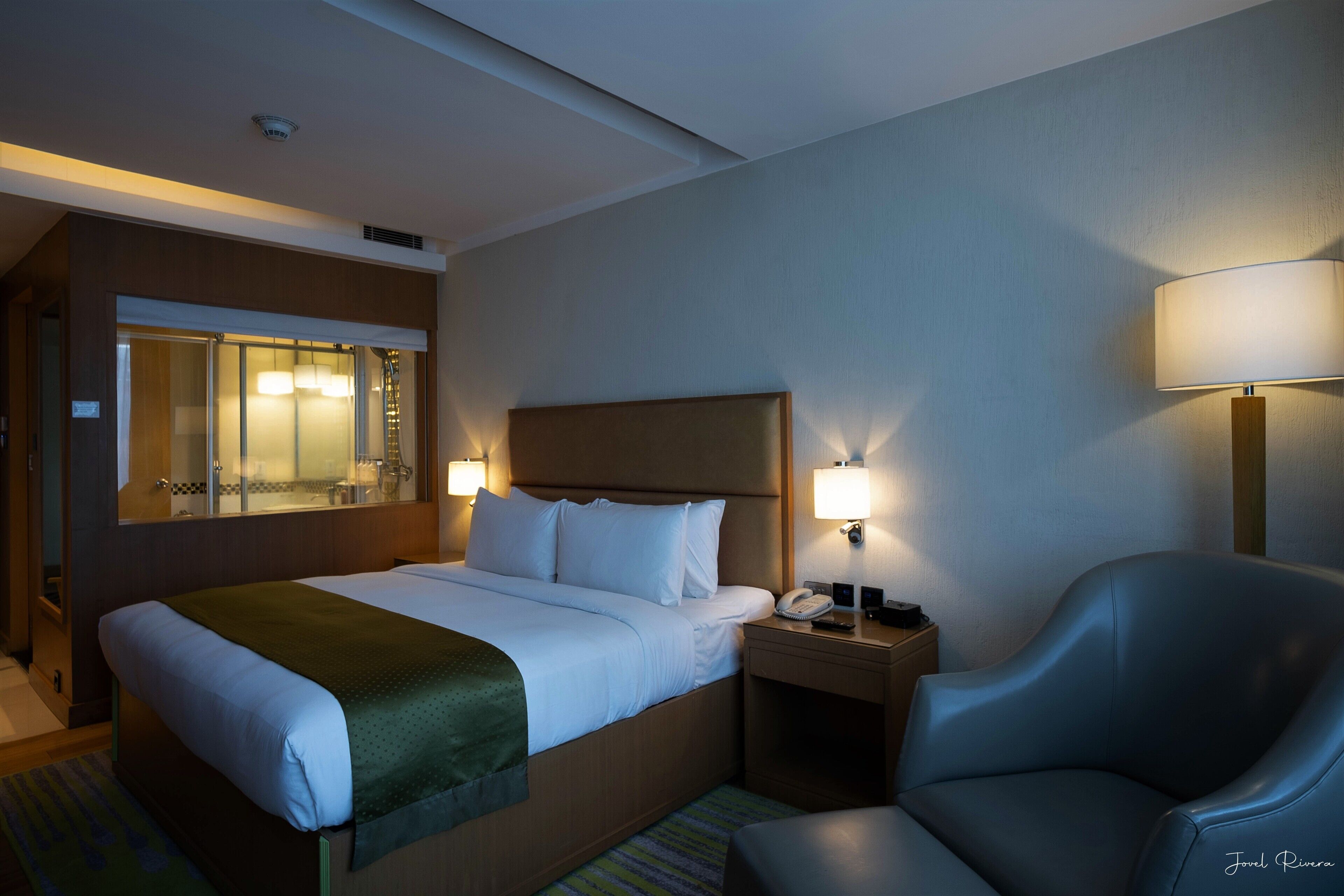 Una habitación de hotel con una cama, lámparas de noche, una silla, una mesita de noche y un espejo.