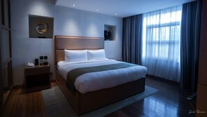 A modern hotel room with a large bed, a bedside table with a phone and a lamp, a wall-mounted circular decoration, and a window with sheer curtains. - Ion Hotel (Baguio)
