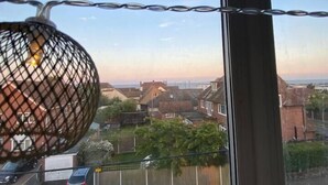 Vista a través de una ventana con una lámpara de malla metálica, cielo despejado y una zona residencial con casas y coches aparcados.