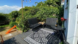Un área de estar al aire libre con un tapete estampado, dos sillas de metal negro, una mesita con una botella y un vaso, y luces colgantes.