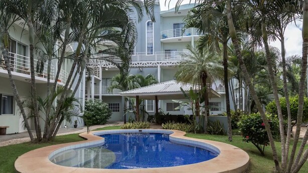 A swimming pool with a surrounding deck, palm trees, and a multi-story building with balconies in the background. - *NEW* Casita Lalita -  2bd/2.5 ba condo - 5 mins to beach w/ 200mbps wifi + pool (Tamarindo)