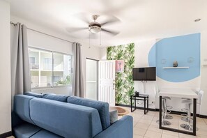 A modern living room with a blue sofa, a ceiling fan, a TV mounted on the wall, and a dining area with a table and chairs.