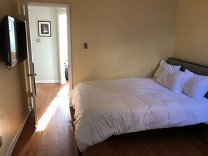 A bedroom with a large bed, wooden flooring, a TV mounted on the wall, and a door leading to another room.