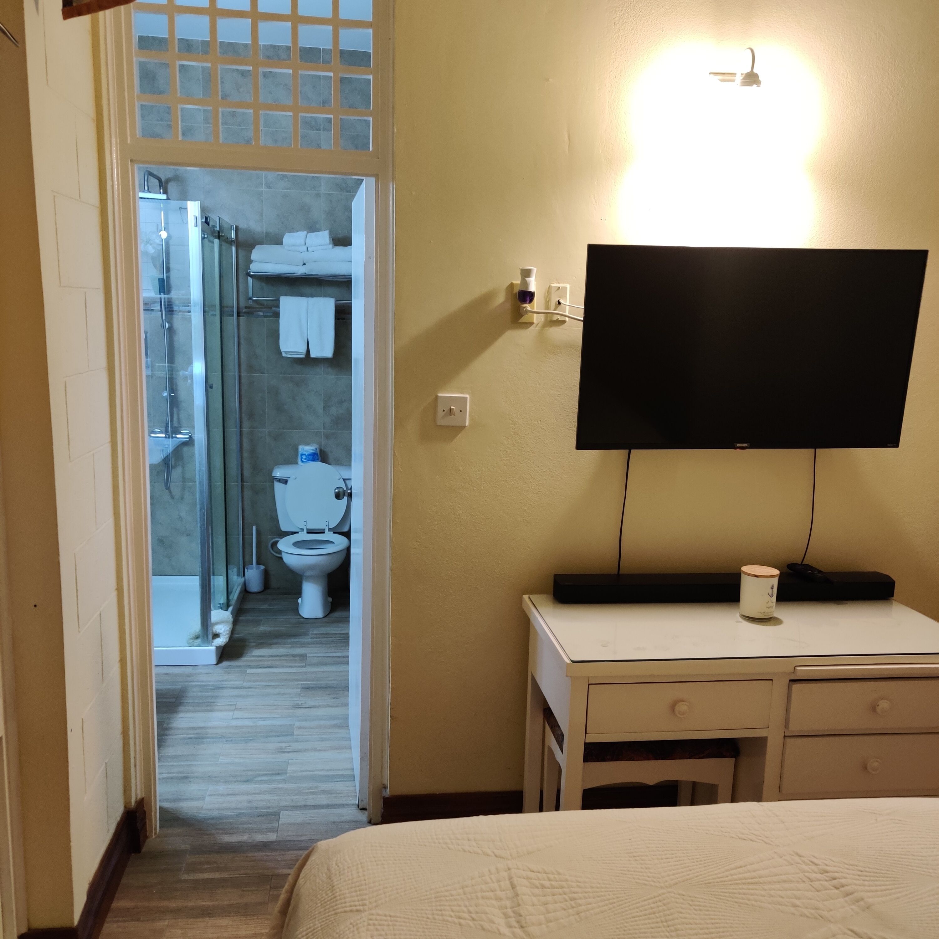 A hotel room with a bed, a desk with a flat-screen TV, a bedside table with a remote control, and a bathroom visible through an open door.