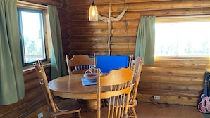 Interior de una cabaña de madera con una mesa redonda para comer rodeada de sillas de madera, una lámpara colgante y una ventana con cortinas.