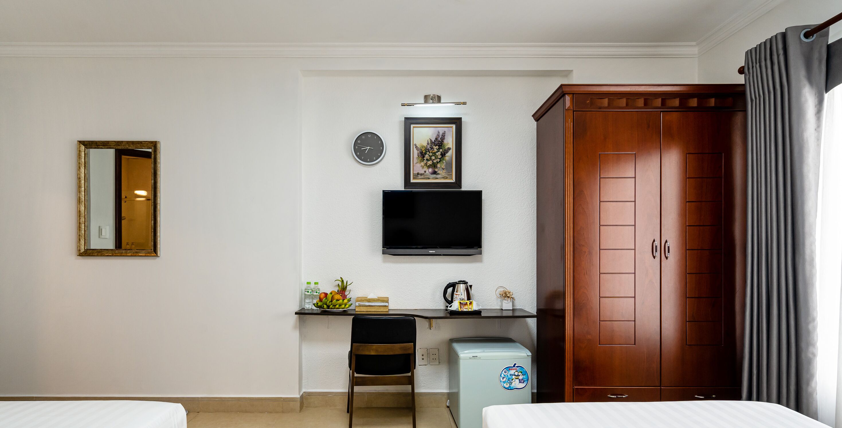 A hotel room with a bed, a desk with a chair, a television, a wardrobe, and a small refrigerator.