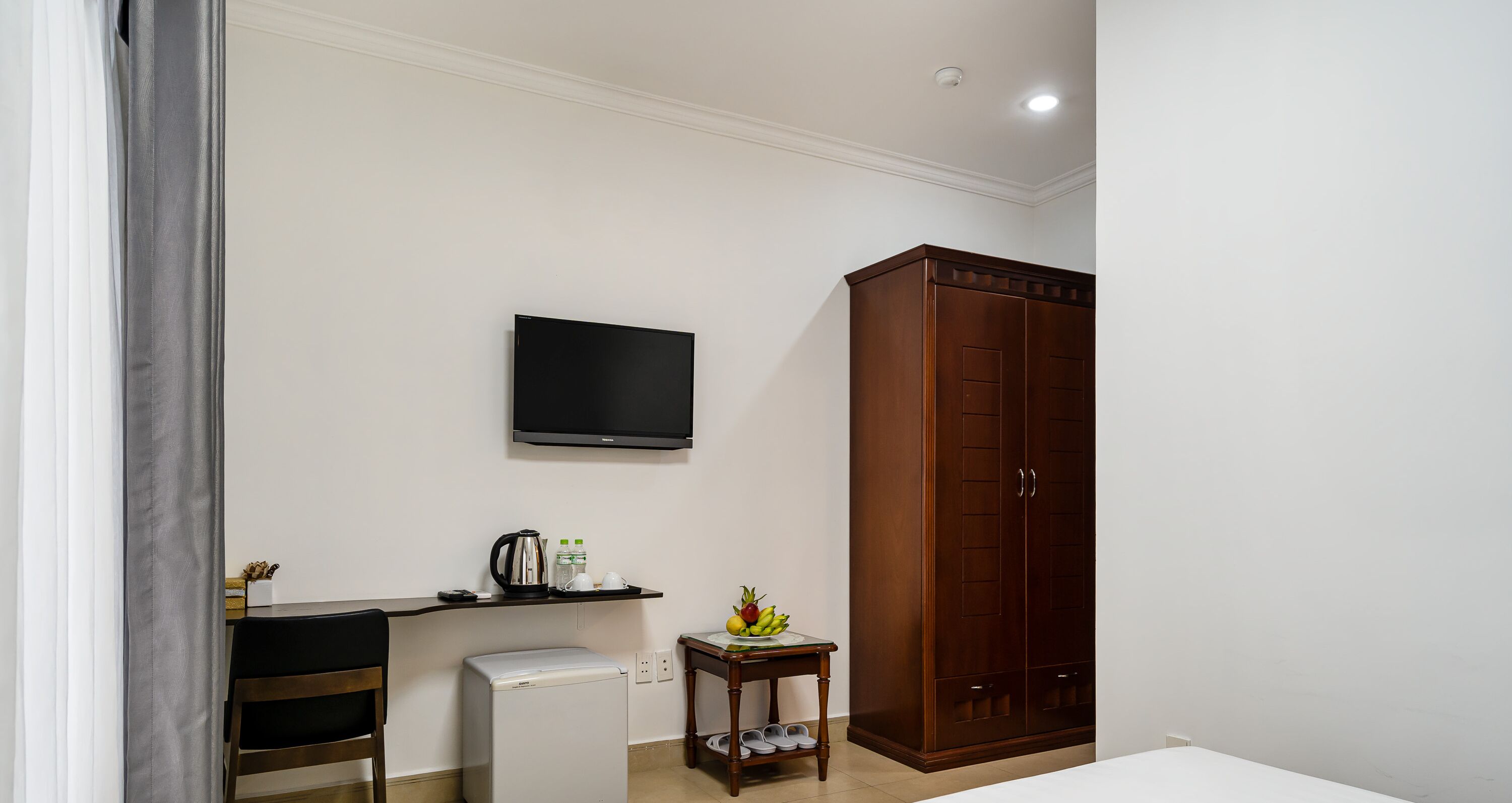 A hotel room with a bed, a desk with a computer, a television, a wardrobe, and a small table with a fruit bowl.