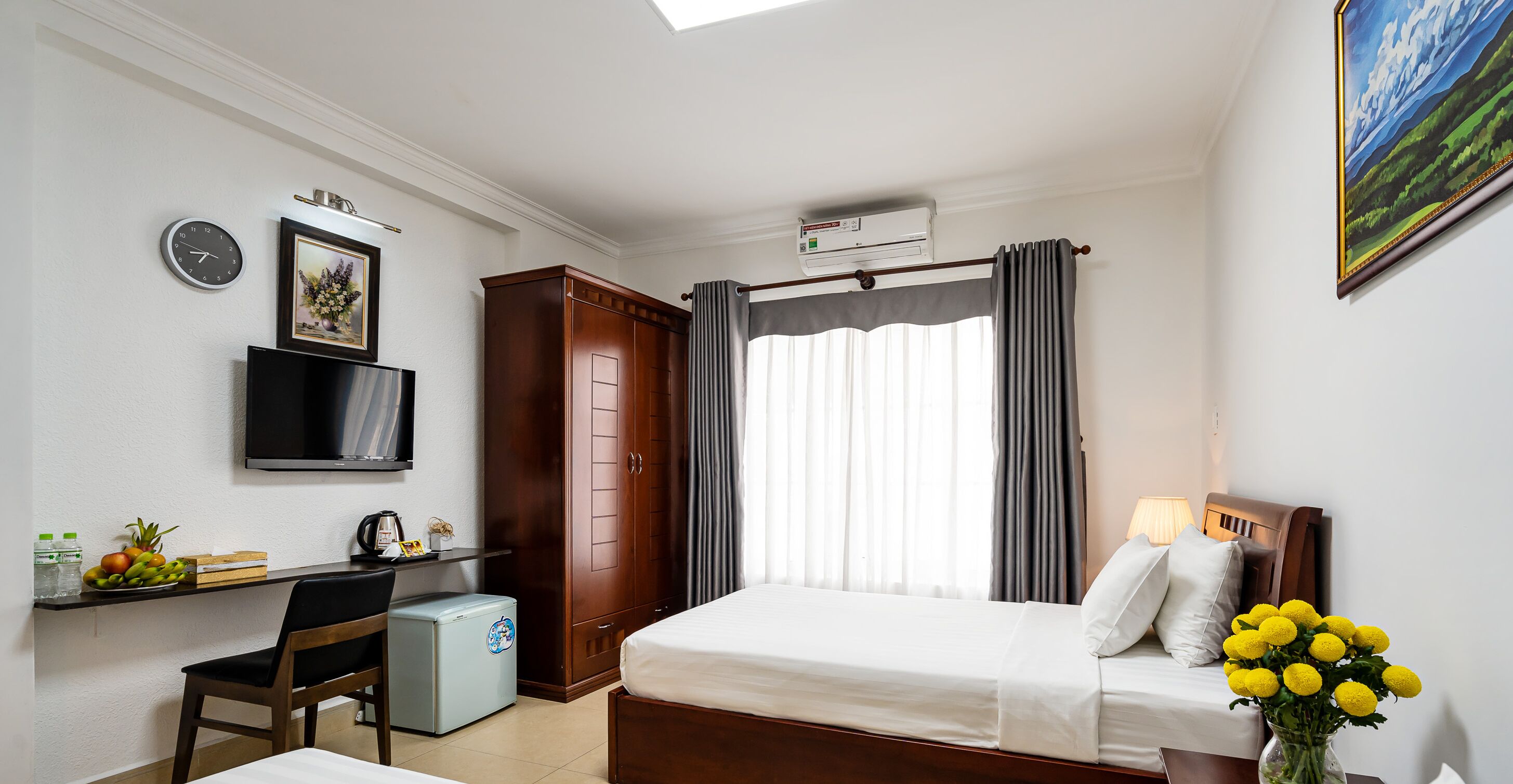 Hotelkamer met een bed, bureau met stoel, kleine koelkast, televisie, wekker en raam met gordijnen.